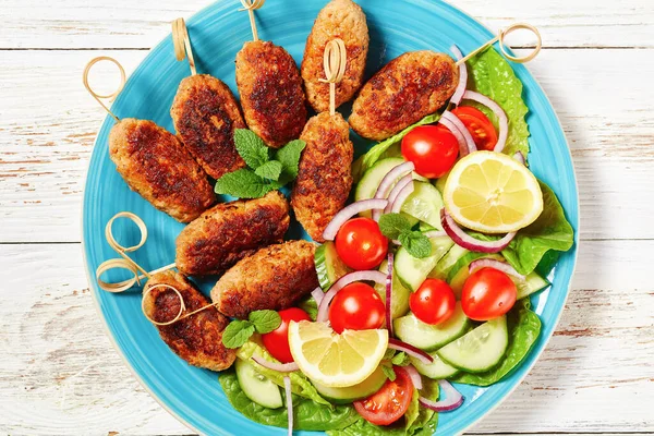 Lamb kofta kebabs of ground lamb, served on a blue plate with yogurt dressing, fresh vegetables salad, and lemon wedges, greek cuisine on a white wooden table, top view, close-up