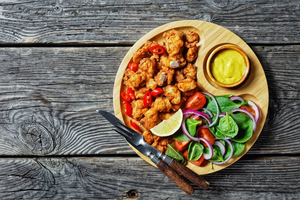Crispy popcorn chicken american fast food served on a bamboo plate with american mustard and salad of baby spinach, red onion, cherry tomatoes and lemon wedge on a wooden table, top view, close-up