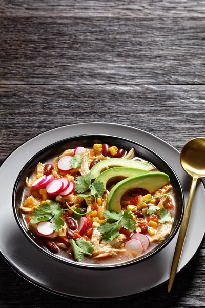 Mexican low carb chicken and tortilla soup with tomato, corn, red kidney beans, avocado, radish and lime, chile, fresh cilantro, on a black plate on a dark wooden background, top view, close-up