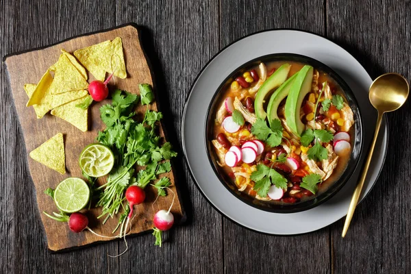 Mexican low carb chicken and tortilla soup with tomato, corn, red kidney beans, avocado, radish and lime, chile, fresh cilantro, on a black plate on a dark wooden background, top view, close-up