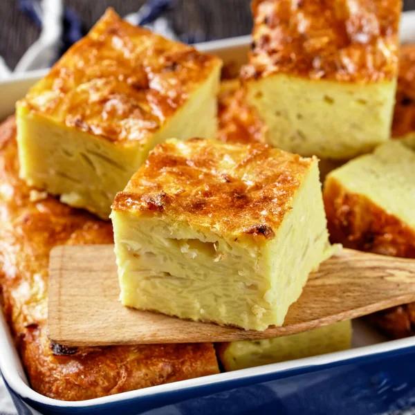 potato kugel, baked pudding or casserole of grated potato cut in portions in a baking dish on a wooden table, jewish holiday recipe, close-up, macro