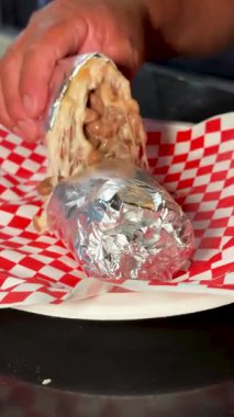 Bir insanın elinde tuttuğu ve folyo kaplı bir burritoyu kareli bir tabakta hafifçe açtığı bir fotoğraf..