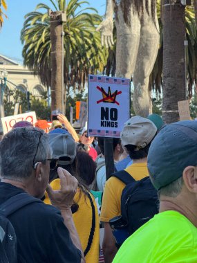 Kalabalık arasında bir protestocu, arka planda görünen şekillendirilmiş bir taç, palmiye ağaçları ve şehir manzarası olan NOKINGS işareti kaldırıyor..