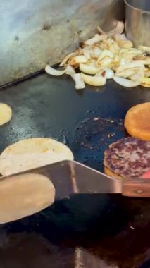 Bir el, bir spatulayı, kızarmış ekmeğin üzerine kızarmış ekmek yapmak için kullanır. Arka planda soğan ve hamburger parçaları vardır..