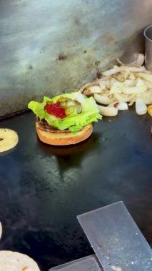 Marul, ketçap ve turşu ile kaplı bir hamburger köftesi ızgara soğan ve çöreğin yanında düz bir ızgaranın üzerinde oturur..