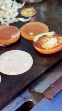 Mısır tortillası ve hamburgerler omuz omuza pişiriliyor. Meşgul bir ızgarada sotelenmiş soğan ile birlikte Latin ve Amerikan sokak yemeklerinin karışımını gösteriyor..