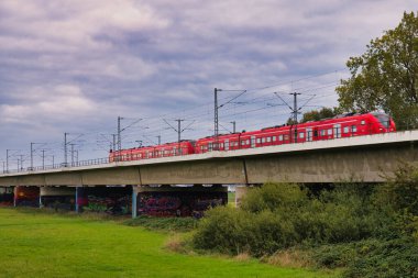 Red commuter train on concrete railway bridge over green field with graffiti underpass and cloudy sky in autumn countryside landscape