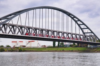 Bulutlu bir günde nehri geçen kırmızı banliyö treniyle modern çelik kemer köprüsü, ulaşım ve kentsel altyapı kavramı