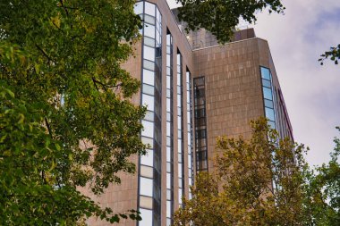 Yeşil ağaçların arkasındaki modern yüksek binalar, şehir merkezinde cam pencereli kentsel mimari, sonbahar mevsiminde doğal ve mimari kontrastlar