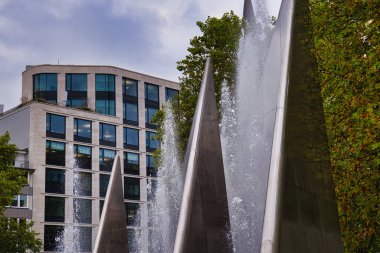Modern mimari arka planı ve şehir merkezinde yeşil yapraklı dinamik su kaynağı.