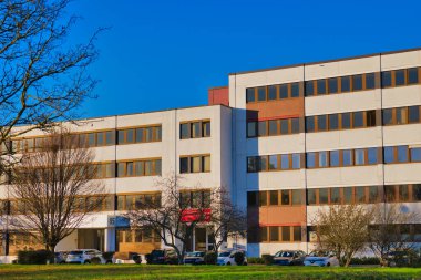 Neuss, Almanya 'da modern bir bina, parlak mavi bir gökyüzünü yansıtan geniş pencereli. Yakınlarda sonbahar ağaçları çıplak, bu da sonbaharın sonları veya kışın başları olduğunu gösteriyor..