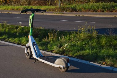 Neuss, Almanya 'da bir yolun kenarında yeşil ve beyaz bir elektrikli scooter duruyor. Güneş manzarayı aydınlatıyor, sonbahar yapraklarını sergiliyor.