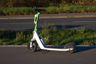 Neuss, Almanya 'da asfalt bir yolun kenarında elektrikli bir scooter duruyor. Yeşil kabzası, motosikletin beyaz çerçevesine karşı parlak bir kontrastlık sağlıyor, yeşillikle çevrili..