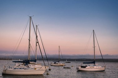 Sakin Marina Sahnesi Durgun Suda Çamurlu Yelkenli Tekneleri Yansıtan Pastel Günbatımı Gökyüzü ve Uzaktaki Sisli Tepeler