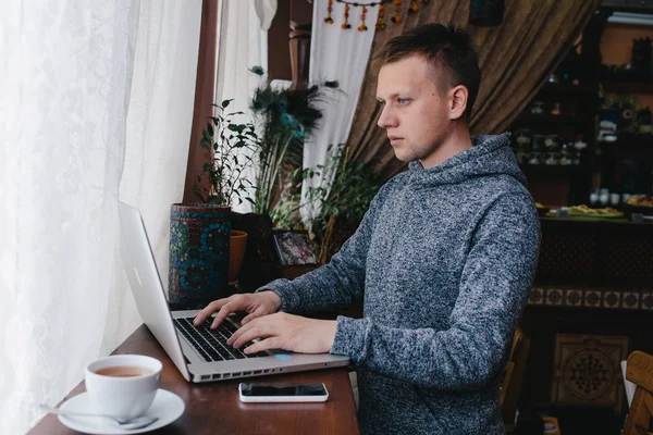 Un hombre usando un portátil y un teléfono celular en un café. Joven bebiendo c — Foto de Stock