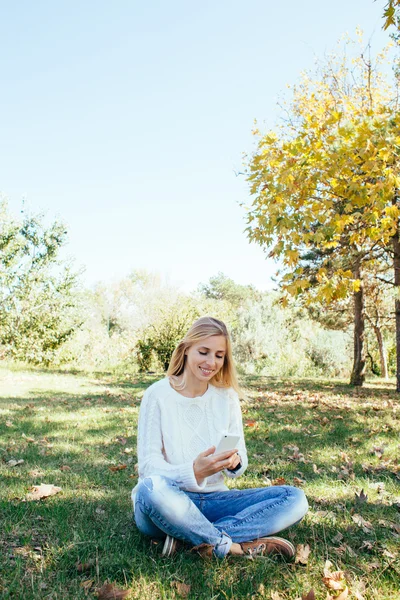 Jovem mulher sentada no parque e usando seu telefone celular. mulher enviando uma mensagem de texto de seu telefone celular ao ar livre — Fotografia de Stock