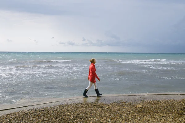 Młoda blondynka w czerwonym płaszczu i ciemnoniebieskich gumowych butach spaceruje po brzegu burzliwego morza — Zdjęcie stockowe