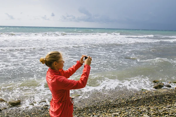 Young woman use mobile phone taking photo on the background stormy sea — Zdjęcie stockowe