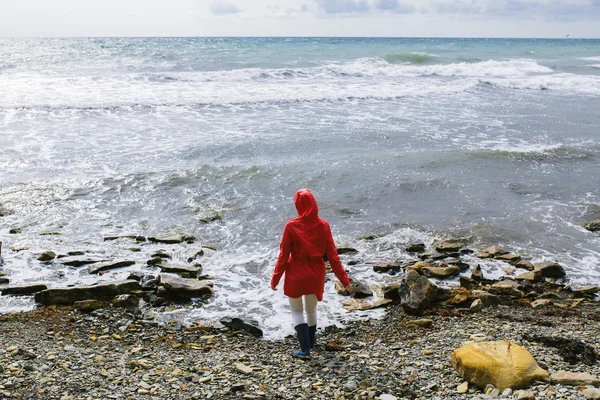 Młoda kobieta w czerwonym płaszczu przeciwdeszczowym i niebieskich gumowych butach wchodzi do wody morskiej — Zdjęcie stockowe