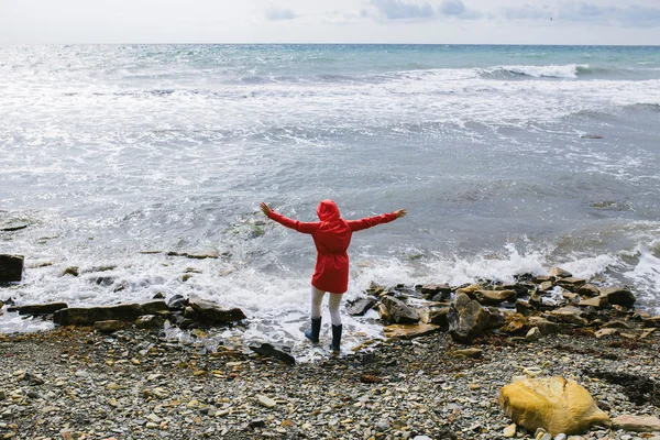 Young woman in a red raincoat and blue rubber boots comes into sea water with raised arms — Stock Photo, Image