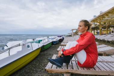 Kırmızı yağmurluklu ve mavi çizmeli genç bir kadın ahşap iskemlede oturuyor ve fırtınalı denize bakıyor.