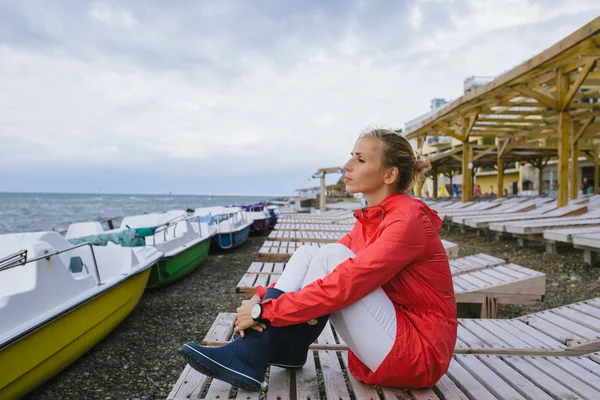Młoda kobieta w czerwonym płaszczu przeciwdeszczowym i niebieskich gumowych butach siedząca na drewnianym leżaku i patrząca na burzliwe morze — Zdjęcie stockowe