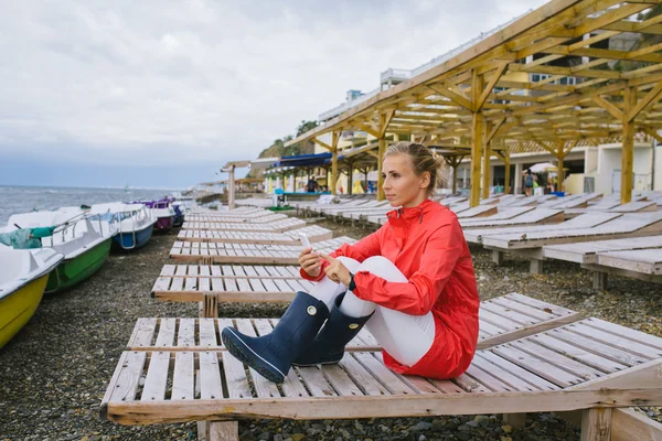 Młoda kobieta w czerwonym płaszczu przeciwdeszczowym i niebieskich gumowych butach, siedząca na drewnianym leżaku nad burzliwym morzem i wysyłająca SMS-a z komórki. Kobieta korzystająca ze smartfona na zewnątrz — Zdjęcie stockowe