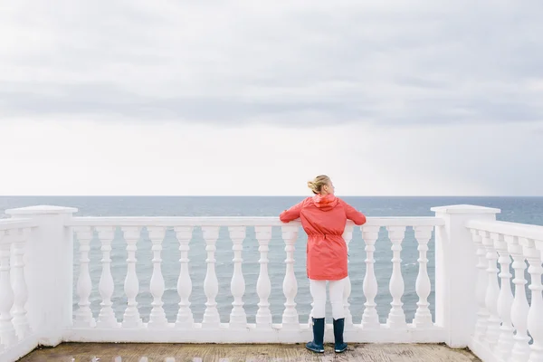 Jeune femme dans une veste rouge et bottes en caoutchouc bleu regarde la mer — Photo