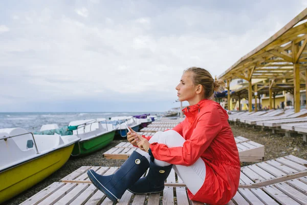 Jeune femme assise sur une chaise longue en bois au bord de la mer orageuse et envoyant un message texte depuis son téléphone portable. Femme utilisant un téléphone intelligent extérieur — Photo