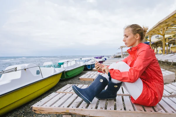 Młoda kobieta siedząca na drewnianym leżaku nad wzburzonym morzem i wysyłająca SMS-a z komórki. Kobieta korzystająca ze smartfona na zewnątrz — Zdjęcie stockowe