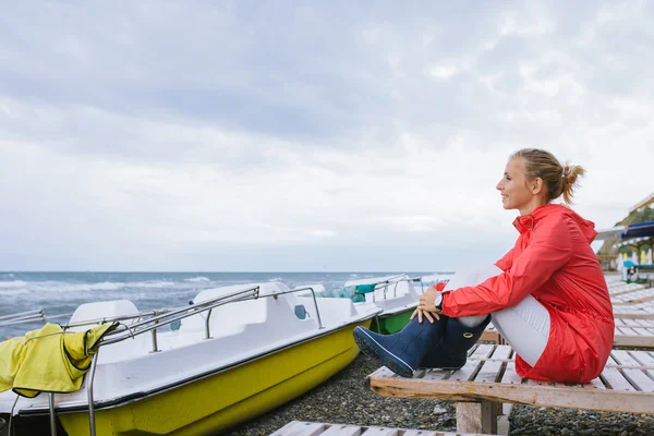 Młoda kobieta w czerwonej kurtce i niebieskich gumowych butach siedząca na drewnianym leżaku i patrząca na burzliwe morze — Zdjęcie stockowe