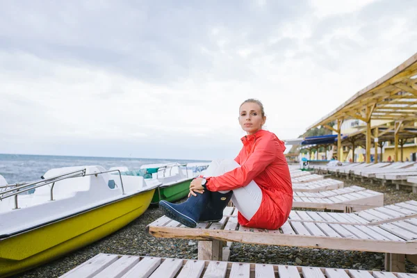 Młoda kobieta w czerwonej kurtce i niebieskich gumowych butach siedząca na drewnianym leżaku w pobliżu morza — Zdjęcie stockowe