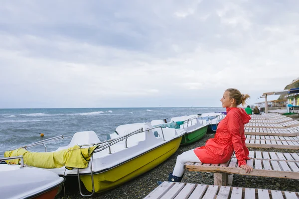 Młoda kobieta w czerwonej kurtce siedząca na drewnianym leżaku i patrząca na burzliwe morze — Zdjęcie stockowe