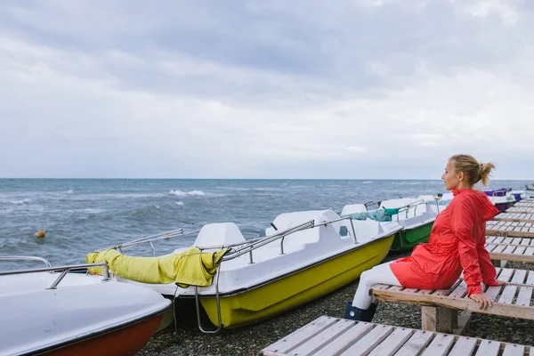 Jeune femme dans une veste rouge assise sur une chaise longue en bois et regardant la mer — Photo