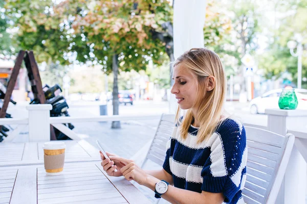 En ung kvinna skickar ett sms från sin mobil på caféet. Kvinna med hjälp av smart telefon på träbordet i uteservering — Stockfoto