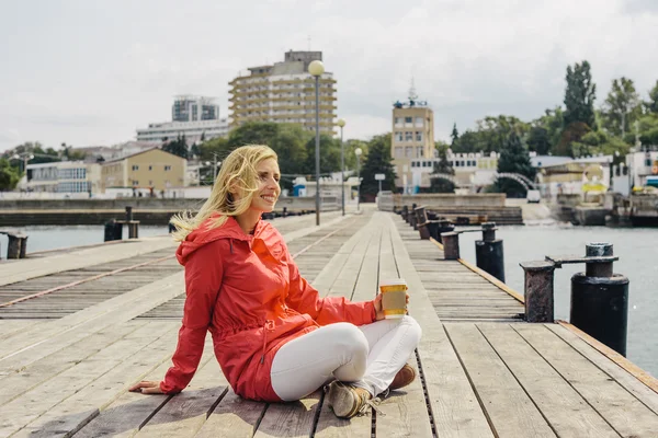 Młoda blondynka w czerwonej kurtce siedzi w doku i trzyma filiżankę kawy — Zdjęcie stockowe
