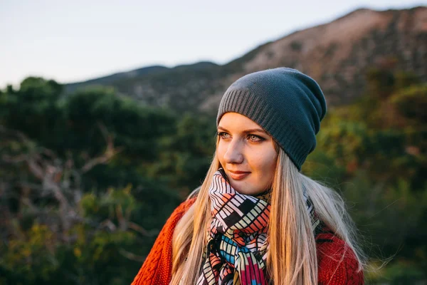 Porträt einer schönen jungen blonden Frau mit Hut, rotem Pullover und buntem Schal im Freien vor dem Hintergrund von Berg und Wald — Stockfoto
