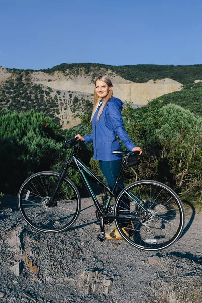 Attraktive junge lächelnde blonde Frau in blauer Jacke steht mit dem Fahrrad und genießt das Leben im Hintergrund Berge — Stockfoto