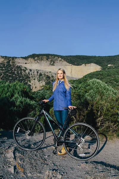 Attraktive junge blonde Frau in blauer Jacke steht mit dem Fahrrad und genießt das Leben im Hintergrund Berge — Stockfoto