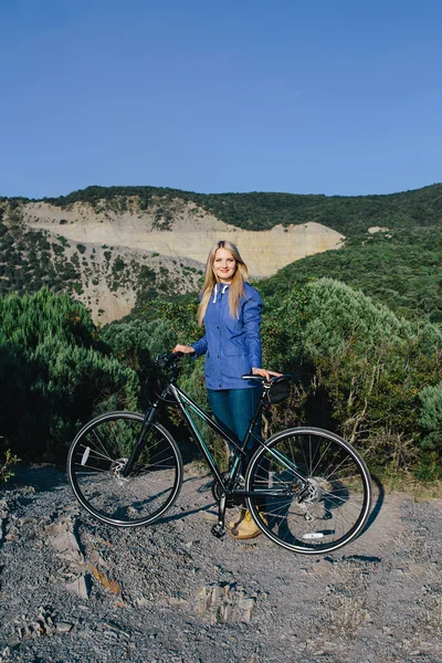 Attraktive junge blonde Frau in blauer Jacke steht mit dem Fahrrad und genießt das Leben im Hintergrund Berge — Stockfoto