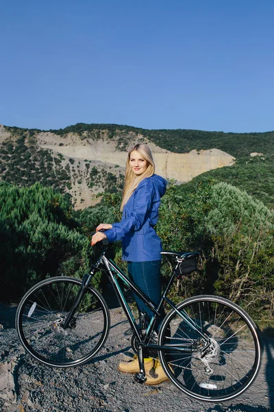 Attraktive junge lächelnde blonde Frau in blauer Jacke steht mit dem Fahrrad und genießt das Leben im Hintergrund Berge — Stockfoto