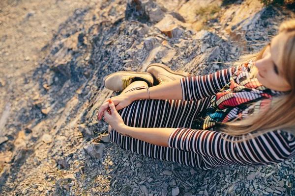 Hermosa mujer rubia joven en un vestido a rayas, bufanda de colores y botas amarillas sentado suelo rocoso al aire libre — Foto de Stock