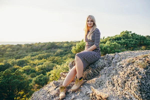 Belle jeune femme blonde dans une robe rayée et bottes jaunes assis sur le fond des forêts — Photo