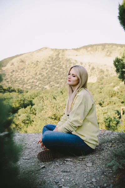 Jeune femme blonde en jeans bleus et bottes de travail jaunes assis en plein air sur un fond de montagnes — Photo