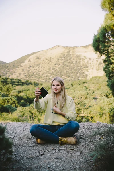 Junge blonde Frau in blauer Jeans und gelben Arbeitsstiefeln, die draußen vor dem Hintergrund der Berge sitzt — Stockfoto