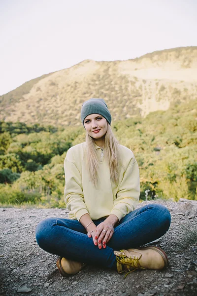 Jeune femme blonde dans un chapeau gris tricoté, jeans bleus et bottes de travail jaunes assis en plein air sur un fond de montagnes — Photo