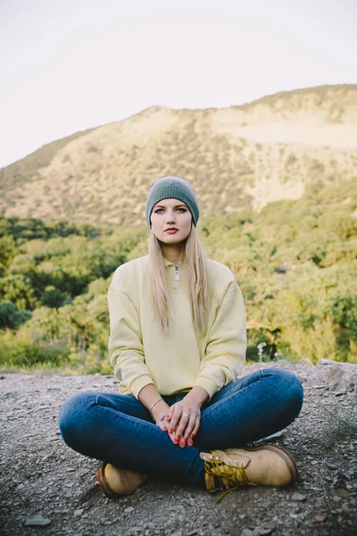 Молодая блондинка в серой вязаной шляпе, синих джинсах и желтых рабочих сапогах сидит на открытом воздухе на фоне гор — стоковое фото