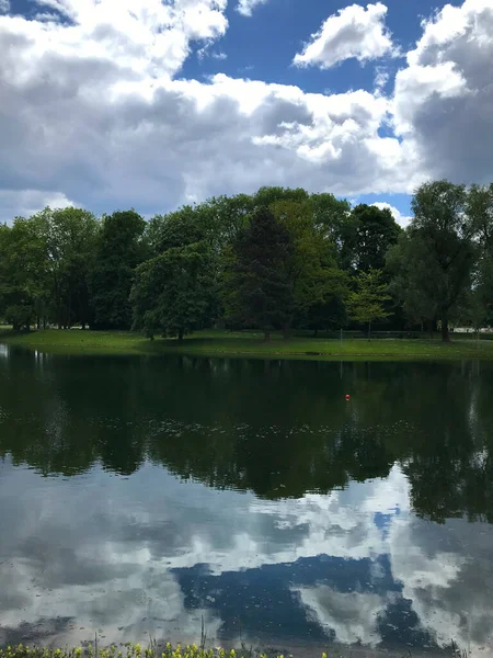 Huzurlu park manzarası, sakin bir göl dramatik beyaz bulutları ve yemyeşil ağaçlarla çevrili mavi gökyüzünü yansıtıyor. Huzurlu su aynası simetrik bir kompozisyon oluşturur, sükunet, denge ve doğal güzelliği taşır..