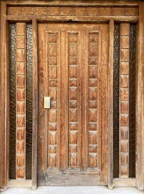 Geometrik oymalı paneller ve desenli kehribar kaplı cam kenarlı klasik ahşap bir giriş kapısı. Simetrik dikey rahatlama detaylarıyla taşralı kahverengi kereste yüzeyi, doğal ahşap tahıl ve klasik mimari çerçeve.