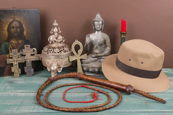 Aventura e conceito arqueológico para artefatos perdidos com chapéu, chicote, vaso de ferro antigo, imagem sagrada, chave da vida, cruzes vintage na mesa de madeira verde e fundo marrom — Fotografia de Stock